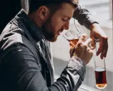 Mężczyzna z brodą, oparty o okno, pije alkohol. W ręku trzyma szklankę z bursztynowym płynem i pustą butelkę. To obraz, jak alkoholik zapije się na śmierć.