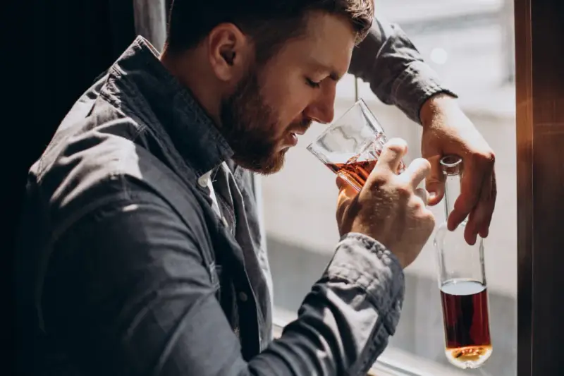 Mężczyzna z brodą, oparty o okno, pije alkohol. W ręku trzyma szklankę z bursztynowym płynem i pustą butelkę. To obraz, jak alkoholik zapije się na śmierć.